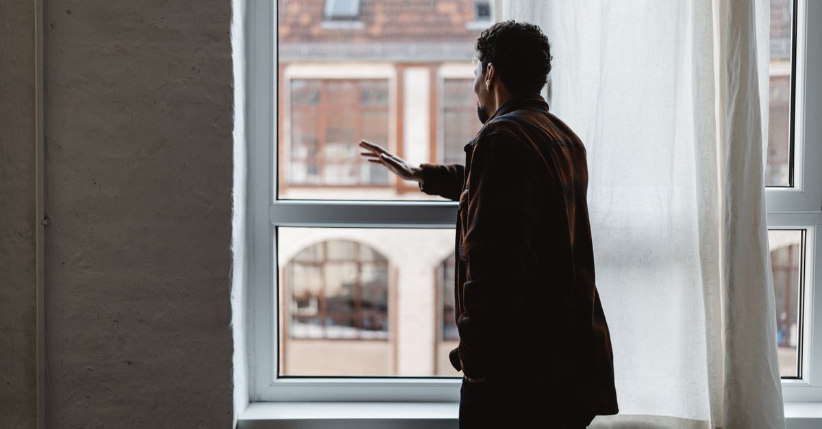 An adult gazing thoughtfully out a large window in a softly lit room.